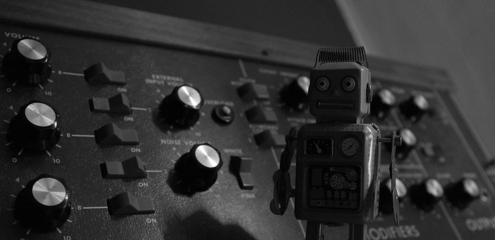 Photo noir et blanc d'un petit robot en métal vintage posé sur un synthétiseur avec de gros boutons.